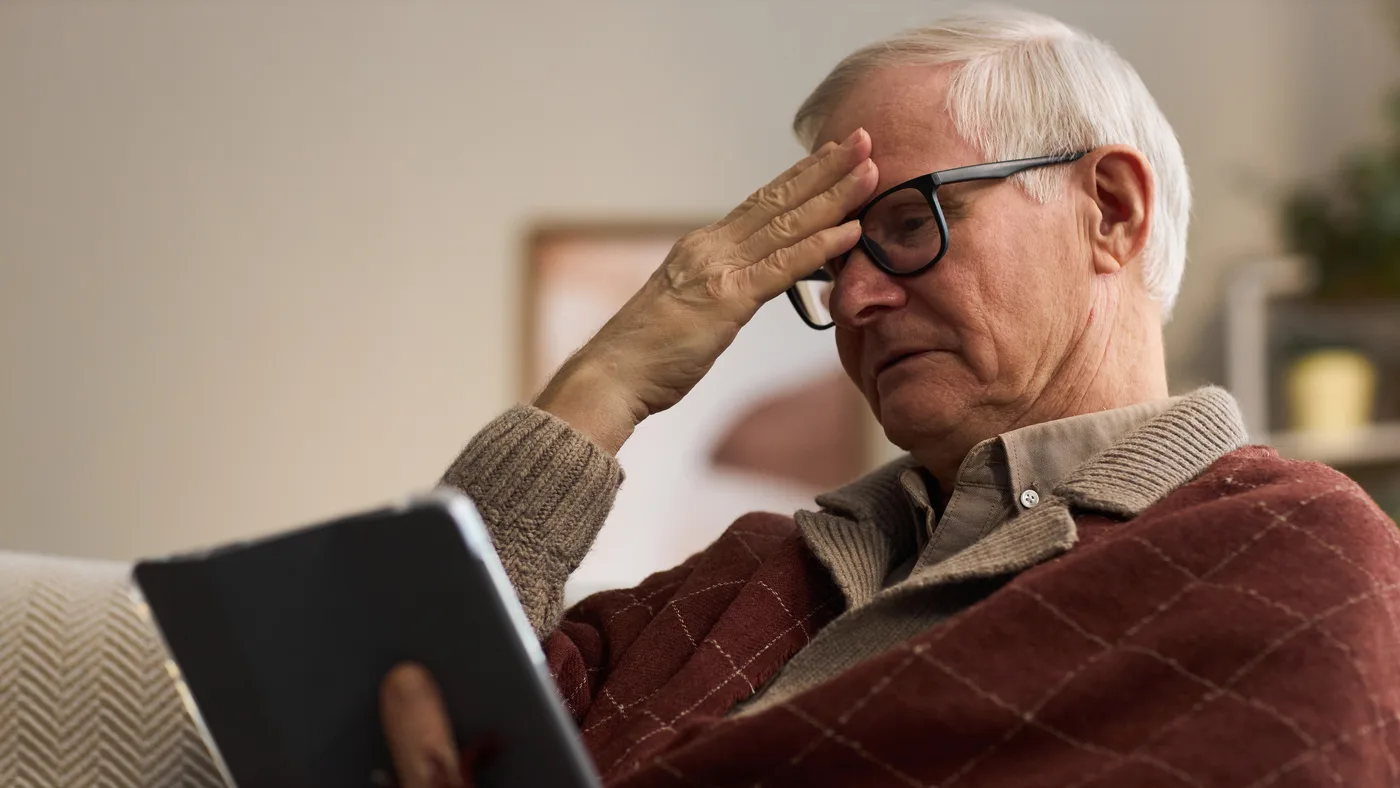 An older man sitting on a sofa with a tablet in his lap, his glasses pushed up and one hand resting against his forehead