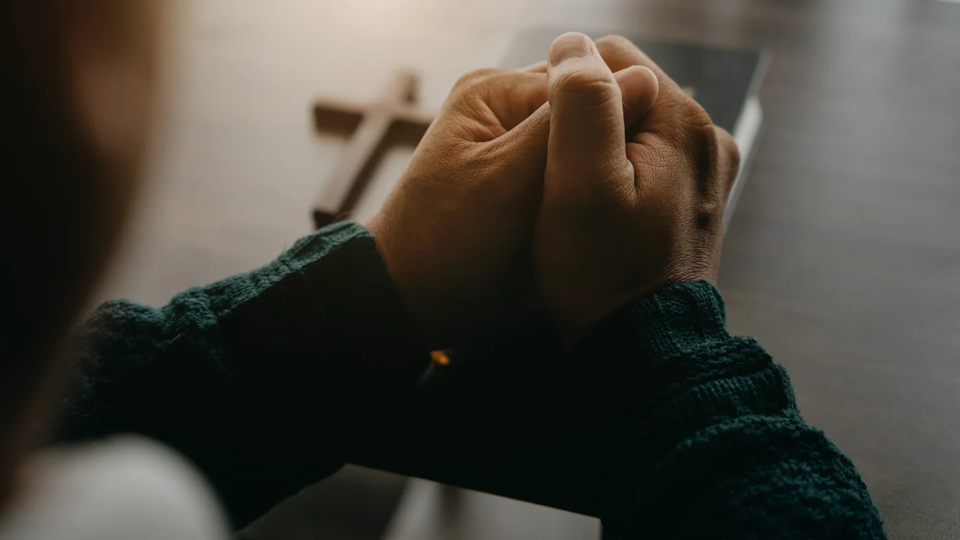 A man's hands folded in prayer on a closed Bible, with a small wooden cross on the table beside them