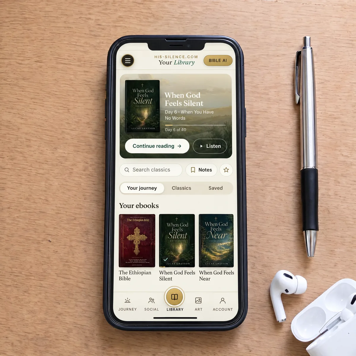 A phone beside wireless earbuds on a wooden desk showing Your Library, the current journey day, the Ethiopian Bible and both journey ebooks